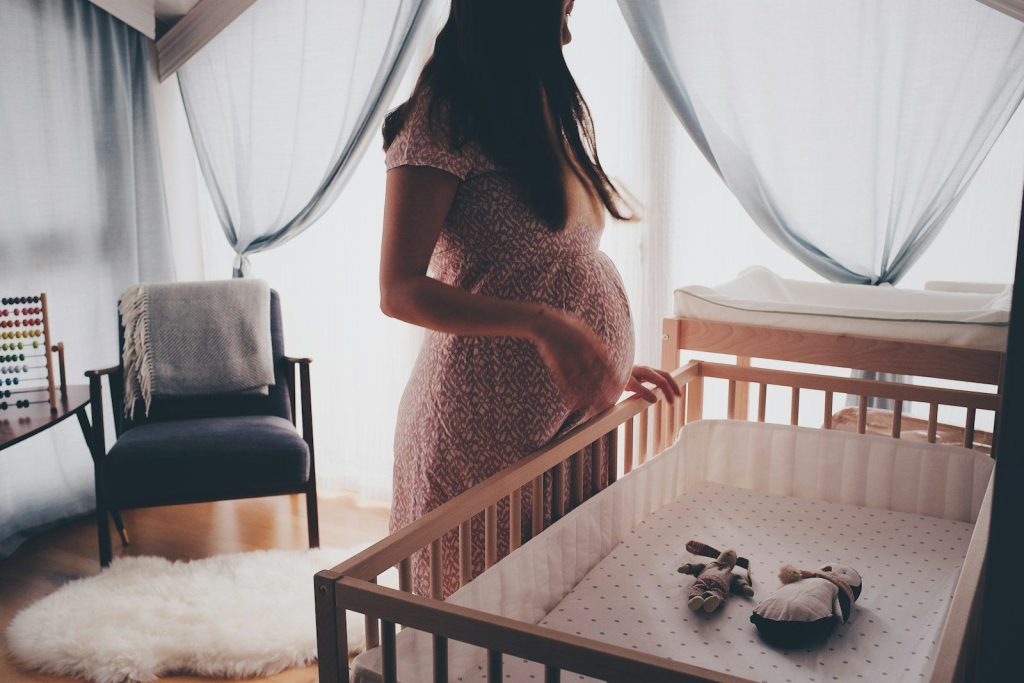 A pregnant woman standing by a wooden crib in a sunlit nursery